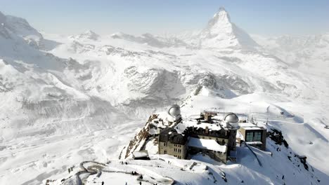 Aerial-flyover-over-Gornergrat-towards-the-Matterhorn-and-descending-train-during-winter-with-snow-and-skiers