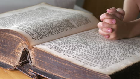 Young-child-reading-bible-with-hands-crossed-on-top-of-book,-close-up-view