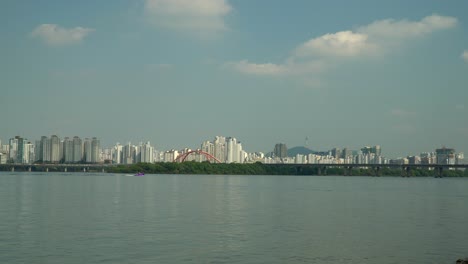 Unrecognizable-Person-Water-Skiing-on-Hangang-River-Distant-view,-Cars-Traffic-on-Seogang-Bridge,-Gangbyeon-Expressway-Road,-Mapo-gu-Apartment-Buildings-and-Seoul-Namsan-Tower-Behind-Bamseom-Island