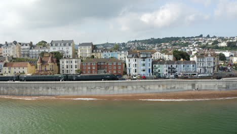 A-profile-drone-shot-of-a-train-along-Dawlish-town-with-seafront-houses-in-the-background-with-the-sea-and-beach-in-shot
