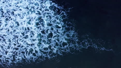Straight-down-aerial-view-of-a-wave-breaking-with-bubbles-and-sea-foam-creating-an-abstract-white-water-pattern-in-the-water