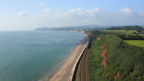 Eine-Luftaufnahme-Eines-Langen-Zuges,-Der-Entlang-Der-Küstenbahn-Von-South-Devon-Fährt,-Mit-Blick-Auf-Das-Blaue-Meer-Und-Die-Küste,-Die-An-Einem-Sonnigen-Sommertag-In-Richtung-Dawlish-Town-Fährt