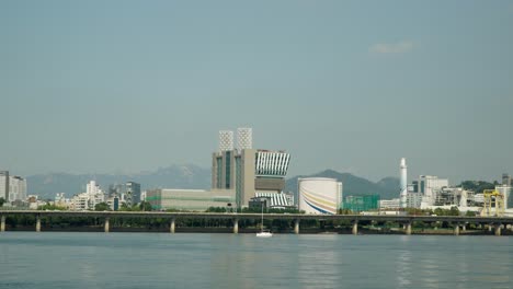 Small-Yacht-with-Folded-Sails-Cruising-on-Hangang-River-with-Gangbyeon-Expressway-Road-and-Mapo-gu-District-of-Downtown-Seoul-in-Background,-Bukhansan-Mountains-behind-the-cityscape