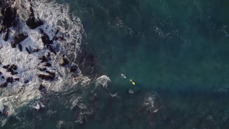 Two-surfers-wating-for-waves-near-rocky-coastline,-top-down-zoom-in-drone-view