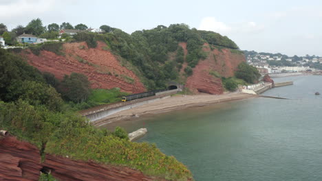 An-aerial-shot-flying-over-red-cliffs-at-Dawlish-with-a-train-coming-out-of-a-tunnel-on-the-Devon-coast-with-the-beach-and-sea-in-shot