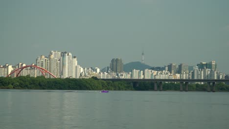 Unrecognizable-Person-Fall-Down-while-Water-Skiing-on-Hangang-River-pulled-by-Speedboat-Distant-view,-Cars-Traffic-on-Seogang-Bridge,-Apartment-Buildings-and-Seoul-Namsan-Tower-Behind-Bamseom-Island