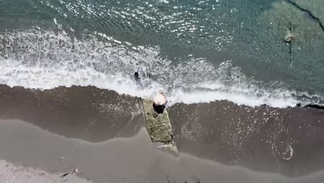 aerial-shot-of-the-shore-of-the-beach-in-ocoa,-young-boy-with-big-hat-sitting-enjoying-the-gentle-waves-of-the-sea