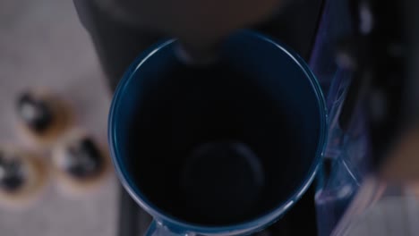 Hand-Puts-Blue-Cup-By-Modern-Coffee-Machine-and-Pours-Coffee,-With-Capsules-In-The-Background,-High-Angle-Close-Up-With-Rack-Focus