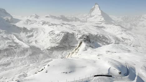 Aerial-flyover-over-Gornergrat-during-winter-with-a-view-of-the-Matterhorn-and-a-descending-train