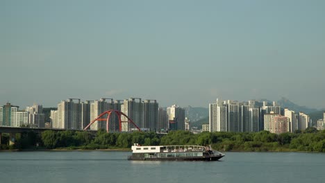 Ferry-Cruise-Ship-Traveling-on-Han-River-along-Bamseom-Island-with-Seoul-Urban-High-Raised-Apartment-Buildings,-Seogang-Bridge-and-Bukhansan-Mountains-on-Background,-tourism-in-South-Korea-Autumn