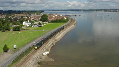 Aerial-shot-of-drone-following-a-train-along-the-River-Exe-approaching-Starcross-station,-on-a-sunny-summers-day