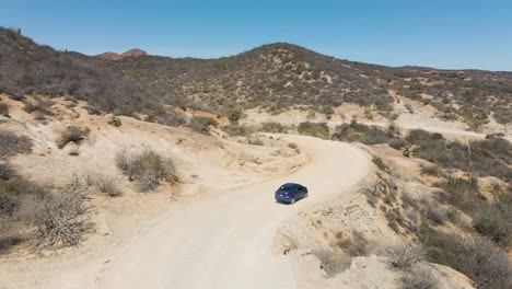 Aerial:-tracking-shot-of-car-driving-on-winding-dirt-road-in-mountain-desert