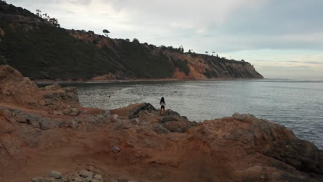 A-woman-stands-on-the-beach-near-Rancho-Palos-Verdes-watching-the-surfers-at-sunset---aerial-parallax-orbit