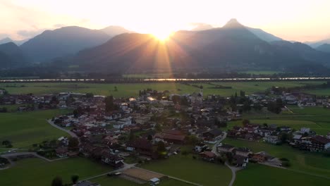 Aerial-view-over-a-summer-forest-during-sunrise-with-mountain-landscape-in-the-Austrian-Alps-with-houses-in-a-small-town-in-the-background