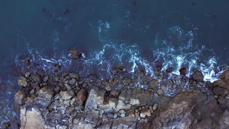 Waves-gently-breaking-against-a-rocky-shoreline---straight-down-aerial-view