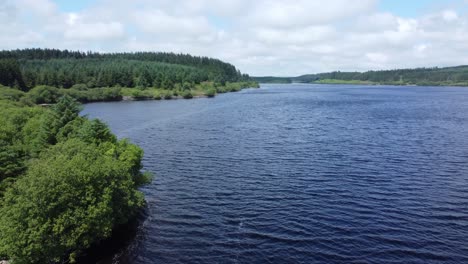 Idyllic-blue-water-reservoir-lake-woodland-hiking-walk-aerial-view-low-orbit-left-reveal-calm-loch