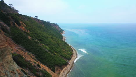 Waves-breaking-against-a-rocky-shoreline-below-the-rugged-terrain-of-Palos-Verdes,-California---pull-back-aerial-view