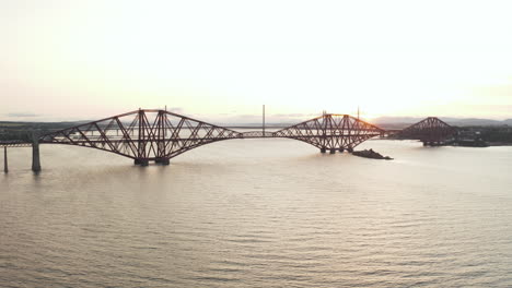A-drone-shot-going-across-the-Forth-Rail-Bridge,-in-Queensferry,-Scotland,-UK-filmed-at-sunset