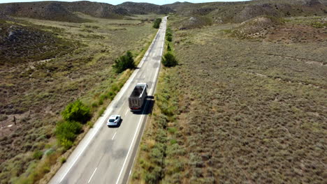 Aerial-view-of-a-Car-On-Road-Trip-Overtaking-Truck-Driving-Along-Desert-Place,-with-windmills-in-the-background