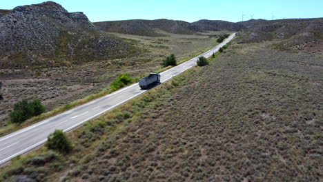 Luftaufnahme-Eines-Lastwagens-Auf-Einem-Roadtrip,-Der-Entlang-Eines-Wüstenortes-Fährt,-Mit-Windmühlen-Im-Hintergrund