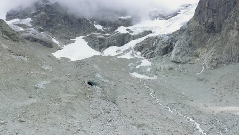Vista-De-Drones-Sobre-Un-Desolado-Glaciar-Alpino-En-Retroceso-Rodeado-De-Montañas-Y-Rocas-Y-Picos-Cubiertos-De-Nieve