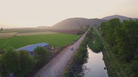 Aerial-shot-as-car-drives-down-rural-road-by-creek-in-between-farmland,-4K
