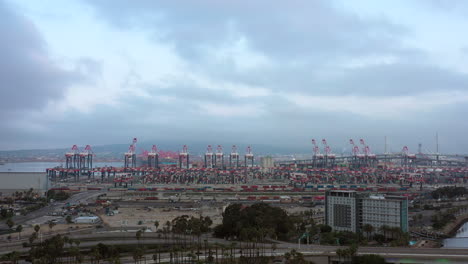 Loading-cranes-and-shipping-containers-at-the-industrial-terminal-port-at-Long-Beach,-California-with-sped-up-city-traffic---aerial-view