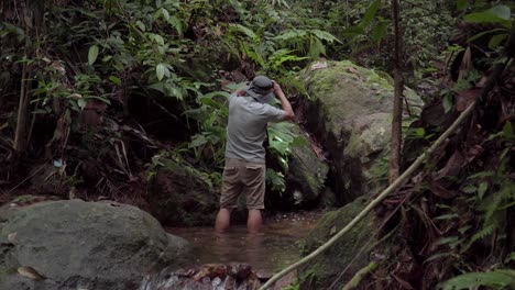 A-male-traveler-hiker-stands-in-a-river,-creek,-stream-looking-around-exploring-and-discovery-with-hiking-headlight-in-jungle,-forest-or-rainforest