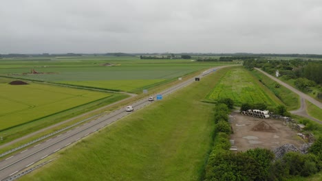 Aerial-tracking-shot-of-traffic-on-a-road-through-agricultural-land-on-the-peninsula-of-Walcheren-in-Zeeland,-the-Netherlands-on-a-cloudy-summer-day