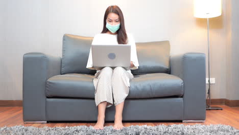 Asian-woman-with-face-mask-sitting-on-sofa-and-typing-on-laptop-computer,-full-frame