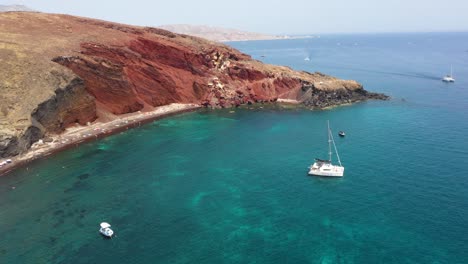 Aerial-spinning-over-Red-Beach-with-turquoise-water,-mountains,-boats-and-red-colored-sand-in-Santorini,-Greece