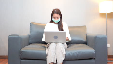 Dressed-in-loose-fitting-clothes-and-wearing-a-surgical-mask,-a-young-woman-works-from-home-on-her-laptop