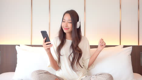Portrait-of-beautiful-exotic-woman-listening-music-from-smartphone-with-wireless-headphones-in-bed,-full-frame-slow-motion