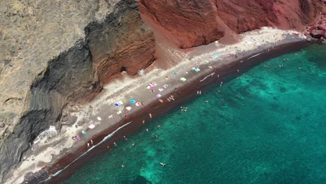 Aerial-top-down-rising-and-revealing-Red-Beach-with-turquoise-water,-tourists,-mountains-and-red-colored-sand-in-Santorini,-Greece