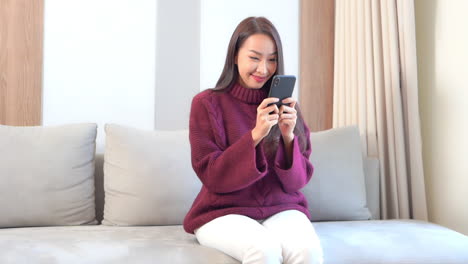 A-pretty-young-woman-in-a-bulky-sweater-and-chinos-sits-on-a-couch-while-she-interacts-with-her-smartphone
