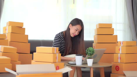 Selling-Online-Concept-Happy-Asian-Woman-Checking-Orders-in-Her-Online-Shop-With-Laptop-With-Room-Full-of-Boxes,-Slow-Motion-Full-Frame