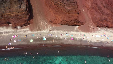 Aerial-top-down-flying-sideways-across-Red-Beach-with-turquoise-water,-tourists,-mountains-and-red-colored-sand-in-Santorini,-Greece