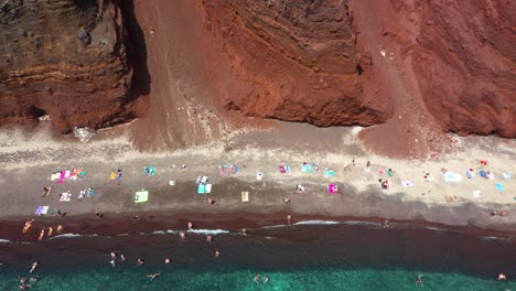 Aerial-top-down-flying-sideways-across-Red-Beach-with-turquoise-water,-tourists,-mountain-and-red-colored-sand-in-Santorini,-Greece
