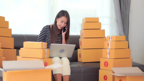 A-young-woman-working-on-her-laptop-is-sitting-among-stacks-of-boxes-waiting-for-shipping-as-she-multitasks-by-answering-her-cell-phone