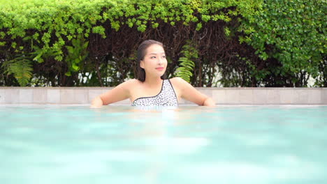 A-young-woman-leans-along-the-side-of-a-resort-pool,-looking-around-at-the-amenities