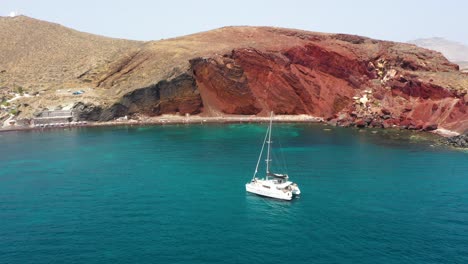 Aerial-descending-over-Red-Beach-with-sailing-boat-passing-by-in-Santorini,-Greece