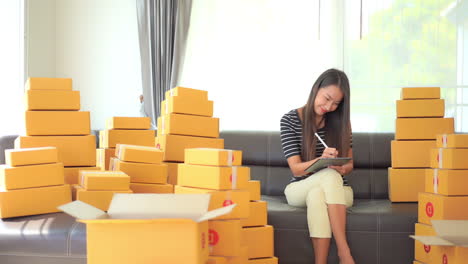 Young-asian-woman,-owner-of-online-shop,-making-list-of-inventory-and-orders-while-sitting-on-sofa-with-product-in-boxes,-full-frame-slow-motion