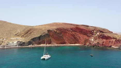 Aerial-flying-sideways-alongside-Red-Beach-with-turquoise-water,-boats,-mountains-and-red-colored-sand-in-Santorini,-Greece