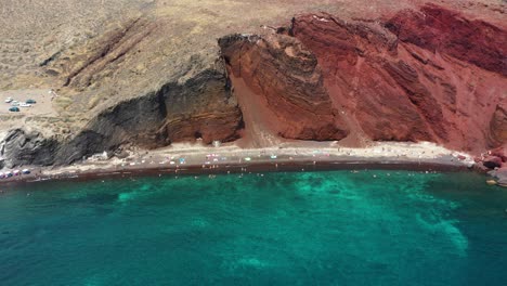 Aerial-drone-flying-towards-Red-Beach-with-turquoise-water,-boats,-mountains-and-red-colored-sand-in-Santorini,-Greece