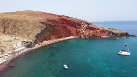 Aerial-drone-revealing-Red-Beach-with-turquoise-water,-boats,-mountains-and-red-colored-sand-in-Santorini,-Greece