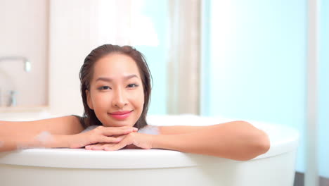 Close-up-of-a-smiling-young-woman-leaning-her-chin-and-arms-along-the-edge-of-a-large-stand-alone-bathtub