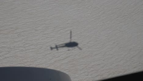 Vista-Desde-Un-Helicóptero-Sobre-Su-Propia-Sombra-En-La-Nieve-Mientras-Volaba-Hacia-Las-Montañas-En-Un-Día-Claro-De-Verano-En-4k