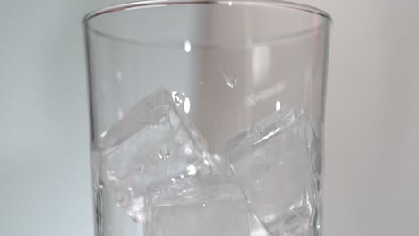 Ice-cubes-in-tall-clear-glass-having-clear-liquid-poured-on-from-metal-jigger-close-up-slow-mo-4k