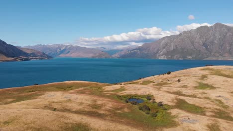 Luftdrohnenaufnahme-Eines-Wunderschönen-Blauen-Sees,-Der-Sich-An-Einem-Sonnigen-Sommertag-In-4k-Hinter-Den-Grasbewachsenen-Hügeln-In-Neuseeland-Versteckt
