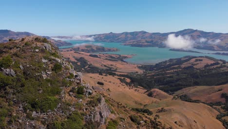 Panorama-Drohnenflug-über-Grasbewachsenen-Hügel-Mit-Leerer-Asphaltstraße-In-Neuseelands-Landschaft,-Vorbei-An-Hohen-Felsformationen,-Feldern-Und-Weiden-An-Einem-Sonnigen-Sommertag-In-4k